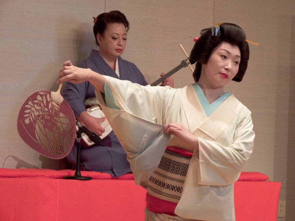 Ms. Shiori,&nbsp;summer fan in her hand, is posing in motion. Ms. Madoka is playing Shamisen in the background