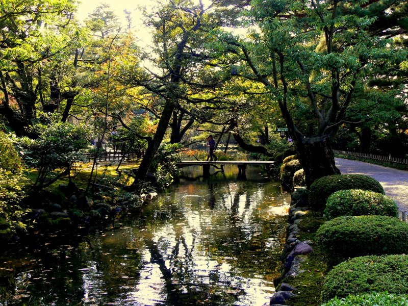 <p>Trees reflected on a pond as a couple walks over one of the garden&#39;s small bridges</p>