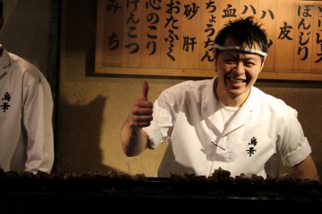 <p>Having some fun cooking in the back, signs behind him show types of yakitori</p>