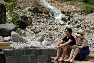 Visitors sit at the edge of a pool with a warm cascade in the background

